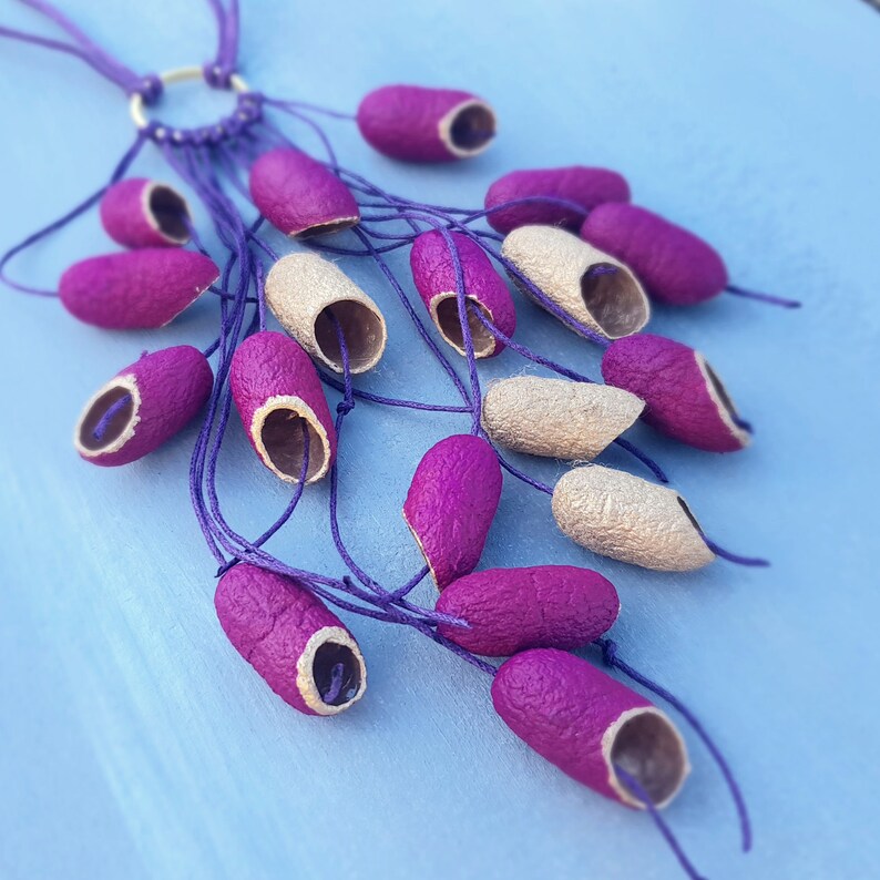 May include: A purple and gold necklace made of strung together seed pods. The necklace is on a blue background.
