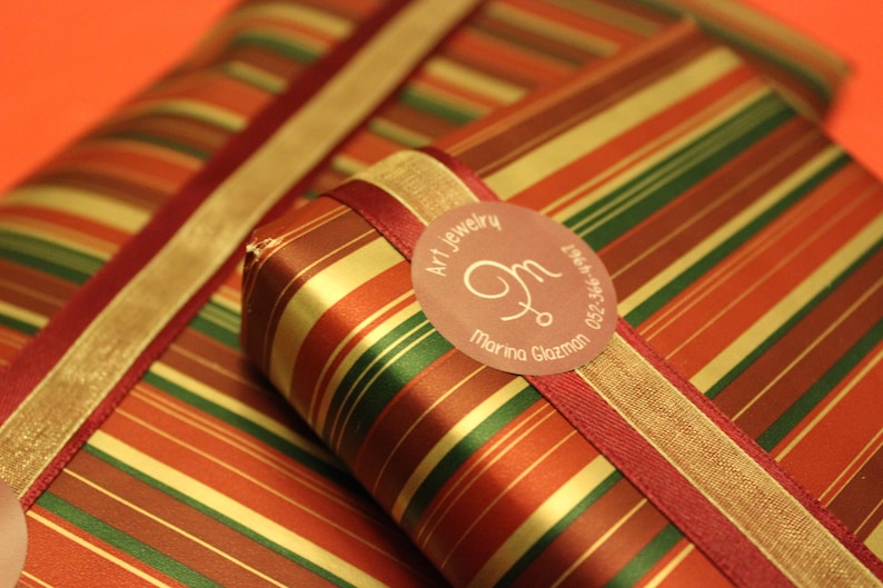 May include: Two red, gold, and green striped gift boxes with a red ribbon and a round, pink label that says "Art Jewelry, M, Marina Glazman, 052-546-2550".