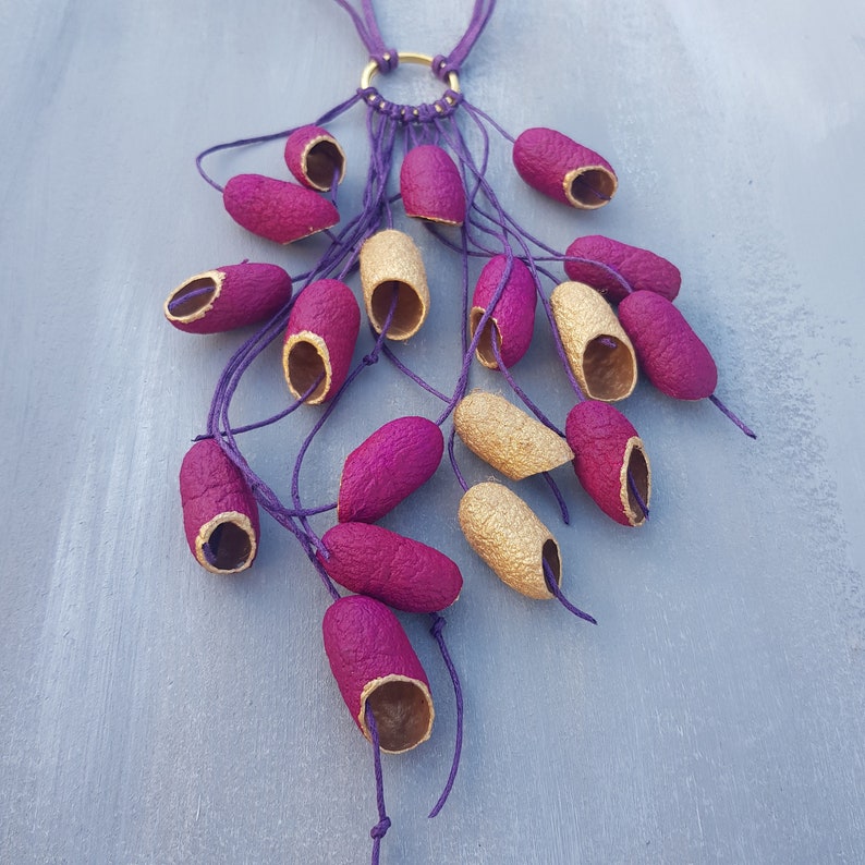 May include: A gold chain necklace with multiple purple and gold seed pods hanging from it. The necklace is made with a macrame style knotting technique.