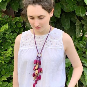 May include: A long, dangling necklace with a purple cord and a gold ring at the top. The necklace is adorned with a cluster of small, red and beige pods.