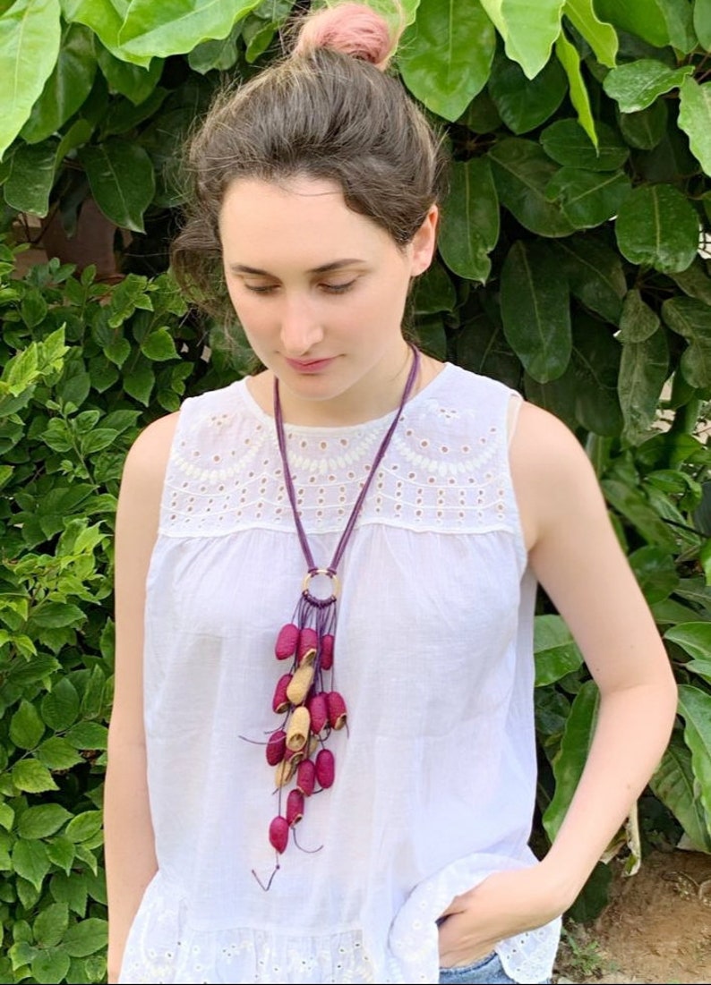 May include: A long, dangling necklace with a purple cord and a gold ring at the top. The necklace is adorned with a cluster of small, red and beige pods.
