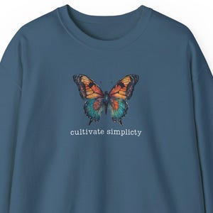 Puede incluir: Una sudadera azul oscuro con un gráfico de mariposa colorida y el texto "cultivate simplicity" impreso en la parte delantera.
