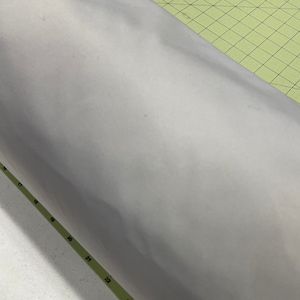 May include: A close-up of a white fabric rolled up on a green cutting mat with black markings. The fabric is smooth and appears to be a lightweight material.