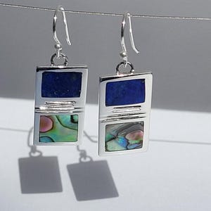 Puede incluir: Un par de pendientes de plata con un diseño de piedras azules y verdes. Los pendientes son de forma rectangular y tienen un cierre simple de gancho.