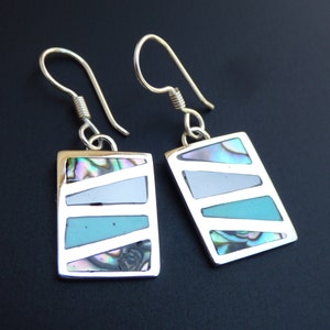 Puede incluir: Un par de pendientes de plata con incrustaciones rectangulares de turquesa y nácar. Los pendientes tienen un diseño simple y geométrico.
