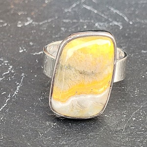 Op de afbeelding: Zilveren ring met een grote, rechthoekige steen. De steen heeft een opvallend patroon van gele, witte en crèmekleurige kleuren. De ringband heeft een getextureerde, gehamerde uitstraling, wat bijdraagt aan het unieke ontwerp.