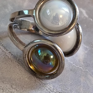 May include: A silver ring with three oval-shaped settings. Two settings hold iridescent, pearly white stones, and the third holds a stone with a rainbow sheen. The ring is set against a gray background.