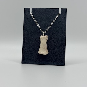 May include: A beige pendant necklace with a silver chain. The pendant is a small, textured, bone-shaped charm. The necklace is displayed on a black stand against a light gray background. The necklace is a piece of jewelry.