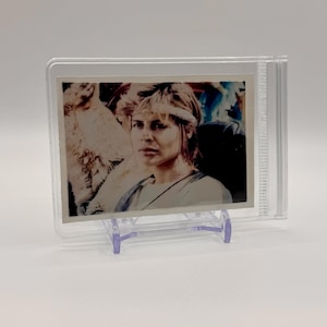 May include: A close-up of a vintage trading card featuring a woman with blonde hair wearing a headband. The card is encased in a clear plastic protector with a stand.