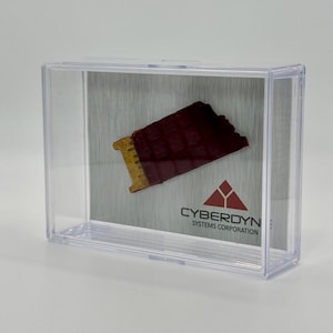 May include: A small, red and gold circuit board fragment encased in a clear plastic display case. The case has a white background with the text "CYBERDYNE SYSTEMS CORPORATION" and a red triangle logo.