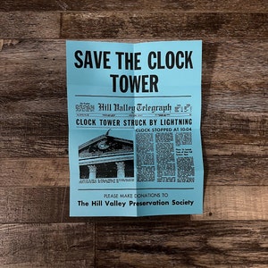 May include: A blue and white poster with the text "SAVE THE CLOCK TOWER" and a newspaper clipping about the clock tower being struck by lightning. The newspaper clipping is from the "Hill Valley Telegraph" and says "CLOCK TOWER STRUCK BY LIGHTNING" and "CLOCK STOPPED AT 10:04". The poster also says "PLEASE MAKE DONATIONS TO The Hill Valley Preservation Society".