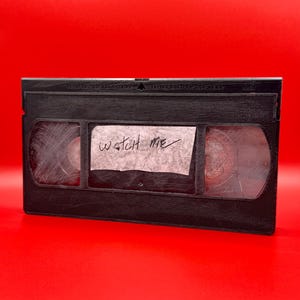 May include: A black VHS video cassette with a white label that says "WATCH ME" written on it.