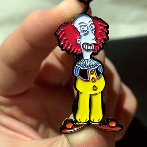 May include: A red and black enamel pin featuring a cartoon character with red hair, a yellow jumpsuit, and a red balloon that says "Die Bart Die".