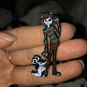 May include: A black and white enamel pin featuring a cartoon version of the character Freddy Krueger from the Nightmare on Elm Street movie series, wearing a striped sweater and a hat. The character is holding the hand of a white cartoon dog.