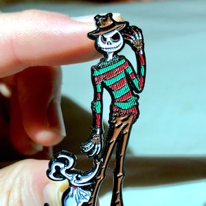 May include: A black and white enamel pin featuring a skeleton wearing a green and red striped shirt and a brown hat. The skeleton is holding a small white ghost with a black bow tie.