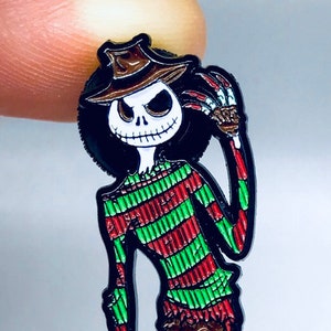 May include: A black and white enamel pin featuring a green and red striped character with a black hat and a white ghost character. The character is holding a knife in one hand and the ghost is standing on its hind legs.