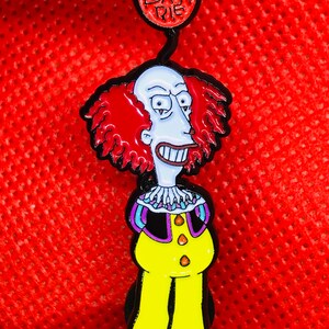May include: A red, white, and blue enamel pin featuring a cartoon clown with red hair and a yellow and black clown costume. The pin has a red balloon attached to the top with the text "Die Bart Die" written on it.