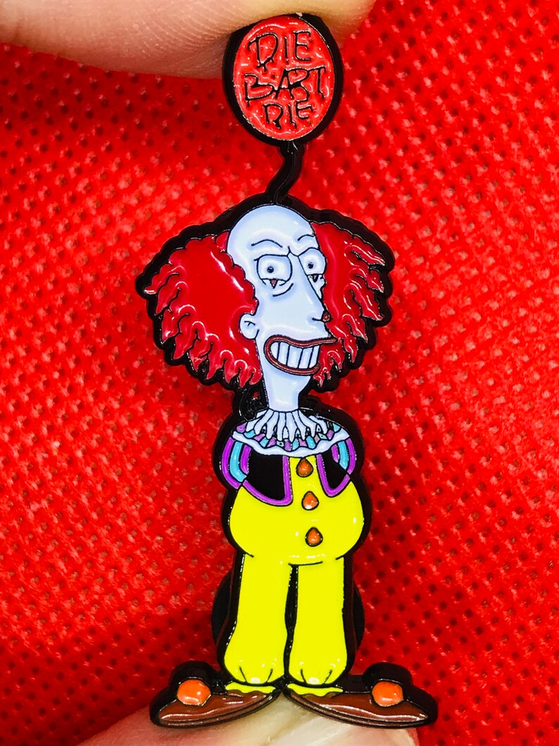 May include: A red, white, and blue enamel pin featuring a cartoon clown with red hair and a yellow and black clown costume. The pin has a red balloon attached to the top with the text "Die Bart Die" written on it.