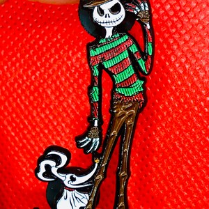 May include: A black and white enamel pin featuring a cartoon version of Jack Skellington from The Nightmare Before Christmas dressed as Freddy Krueger from the Nightmare on Elm Street franchise.  The pin also features a small, white, cartoon dog.