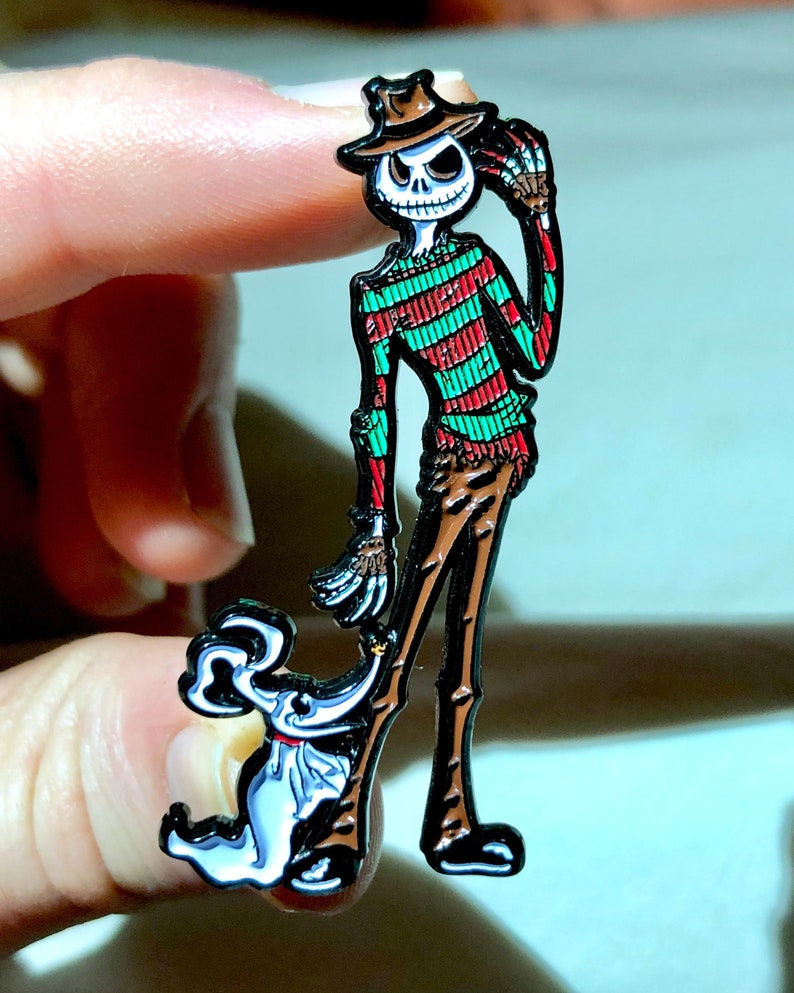 May include: A black and white enamel pin featuring a skeleton wearing a green and red striped shirt and a brown hat. The skeleton is holding a small white ghost with a black bow tie.