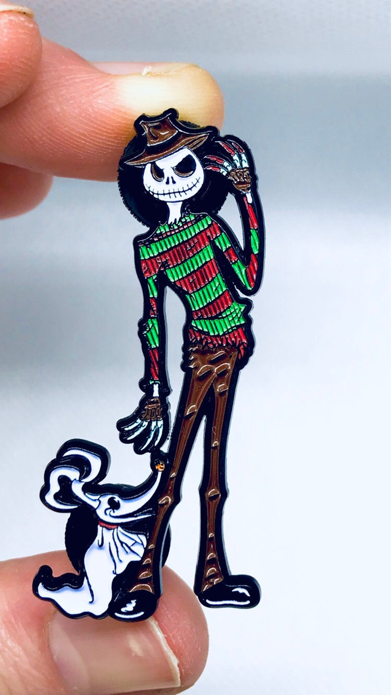 May include: A black and white enamel pin featuring a green and red striped character with a black hat and a white ghost character. The character is holding a knife in one hand and the ghost is standing on its hind legs.