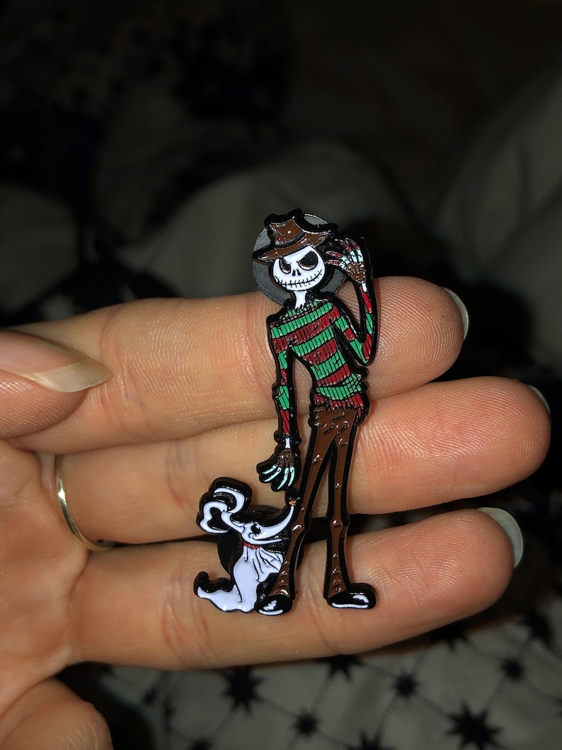 May include: A black and white enamel pin featuring a cartoon version of the character Freddy Krueger from the Nightmare on Elm Street movie series, wearing a striped sweater and a hat. The character is holding the hand of a white cartoon dog.