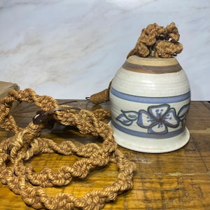 May include: A white ceramic bell with a blue and brown floral design and a brown rope hanging from the top.