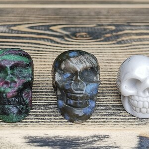 May include: Three carved skull figurines made of different types of stone. The first skull is green and black with pink accents. The second skull is blue, black, and white. The third skull is white.