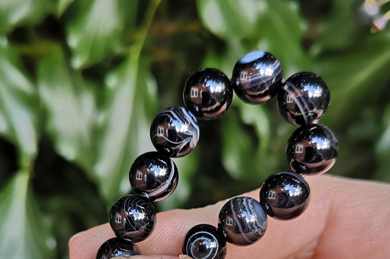 May include: A black and white striped bead bracelet. The beads are round and polished, with a glossy finish. The bracelet is strung together, and the beads have a marbled appearance. The background is blurred green foliage.