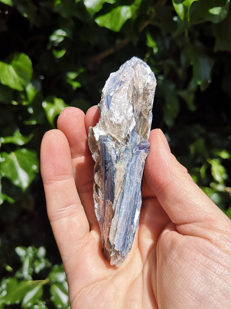K&ouml;nnte beinhalten: Ein blauer Kyanit-Kristall mit silbernem Schimmer. Der Kristall ist roh und hat eine raue Textur. Er ist etwa 10 cm lang und 2,5 cm breit.