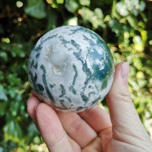 May include: A green and white moss agate sphere, polished to a smooth finish. The sphere is about the size of a golf ball and has a swirling pattern of green and white.
