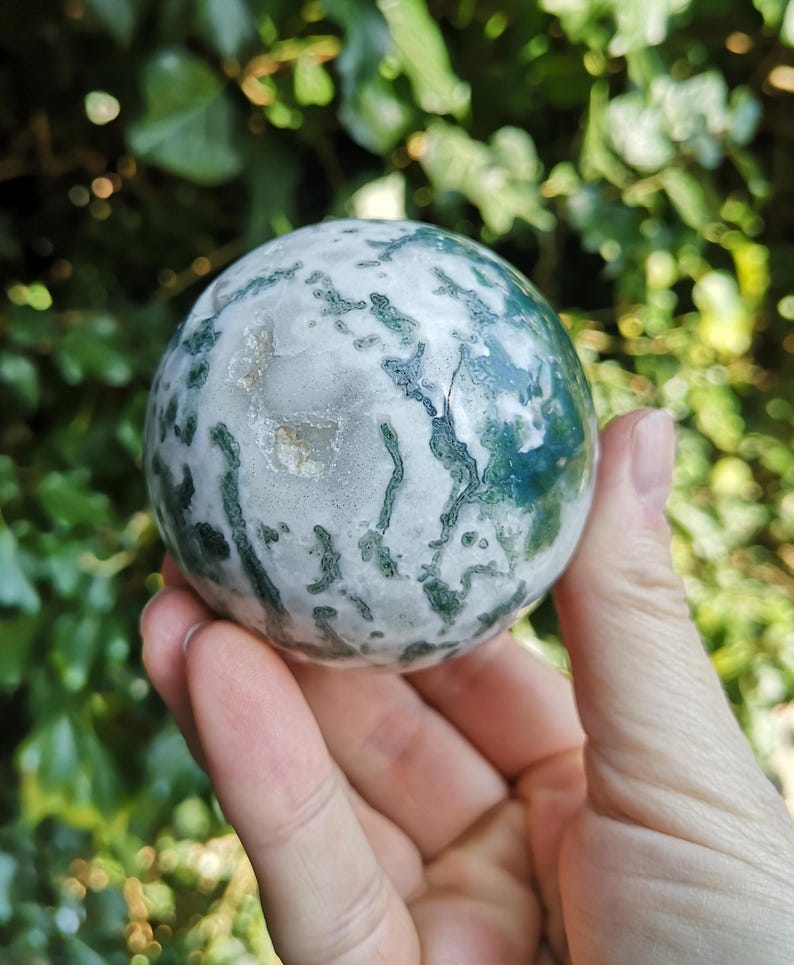 May include: A green and white moss agate sphere, polished to a smooth finish. The sphere is about the size of a golf ball and has a swirling pattern of green and white.