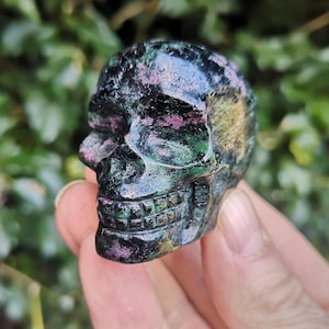 May include: A green, black and pink carved skull made of gemstone. The skull is detailed with a carved mouth and eye sockets.