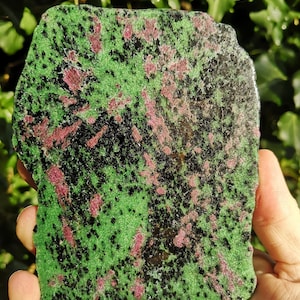 May include: A rough-cut zoisite stone with a vibrant green base, speckled with black and ruby-red inclusions. The natural stone is held in a hand, showcasing its unique, organic shape and color variations. The stone's surface has a slightly textured appearance.