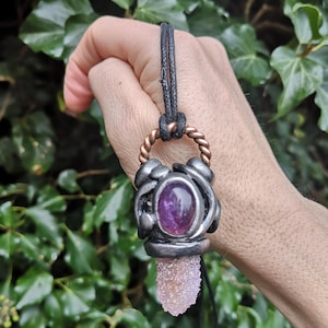 May include: A handcrafted pendant necklace featuring a purple gemstone set in a silver-toned metal. The pendant has a light pink crystal at the bottom and is suspended from a black cord. The necklace is held against a backdrop of green foliage.