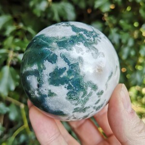 May include: A green and white agate sphere, polished to a smooth finish. The sphere has a swirling pattern of green and white, with a hint of pink in the center.