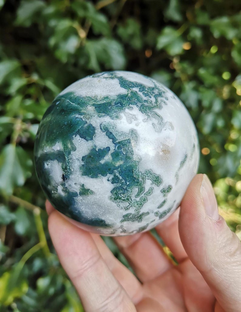 May include: A green and white agate sphere, polished to a smooth finish. The sphere has a swirling pattern of green and white, with a hint of pink in the center.