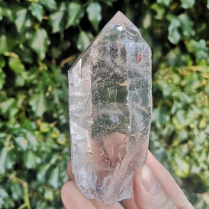 May include: A clear quartz crystal point with a natural, raw finish. The crystal is approximately 12.5 cm tall and has a hexagonal shape. The point is held in a hand, with a green leafy background.