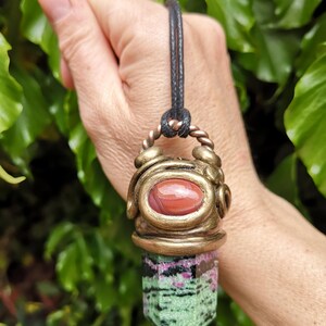 May include: A pendant necklace featuring a gemstone. The pendant has a bronze-colored metal setting with a red oval stone at the top. The lower portion of the pendant is a green and pink crystal, with a clear point at the bottom. The necklace has a black cord.