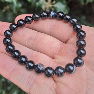 May include: A black beaded bracelet with a mix of solid and patterned beads. The bracelet is made of polished, round beads, strung together to form a flexible band. The beads have a glossy finish, reflecting light. The bracelet is shown on a hand.