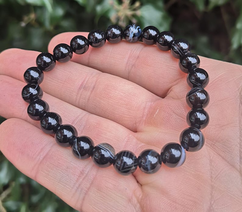 May include: A black beaded bracelet with a mix of solid and patterned beads. The bracelet is made of polished, round beads, strung together to form a flexible band. The beads have a glossy finish, reflecting light. The bracelet is shown on a hand.