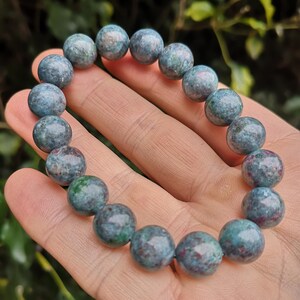 Peut inclure: Un bracelet de perles rondes polies, composées de teintes de teal, vert et marron. Les perles, d'environ 1,3 cm de diamètre, sont enfilées sur un élastique. Le bracelet est présenté sur un fond vert naturel, mettant en valeur les couleurs.