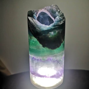 May include: A green, purple, and white fluorite crystal lamp with a light inside. The crystal is shaped like a cylinder with a hole in the top.