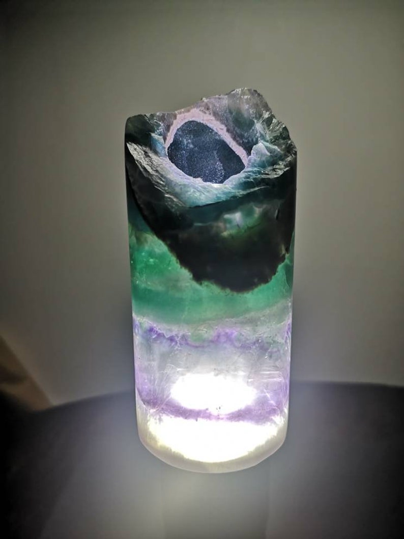 May include: A green, purple, and white fluorite crystal lamp with a light inside. The crystal is shaped like a cylinder with a hole in the top.