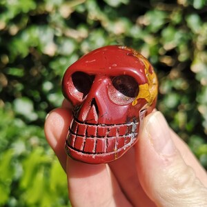 May include: A carved red skull figurine with a detailed jawline and eye sockets. The skull has a polished finish and a small patch of yellow and gold on the top. The figurine is held in a hand against a blurred green background.