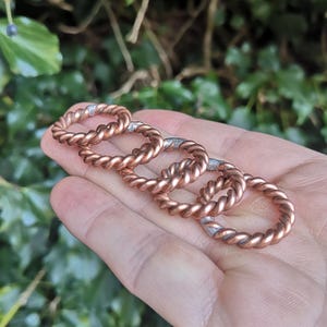 May include: Five copper rings with a twisted rope design. The rings are stacked on top of each other and are held in a hand.