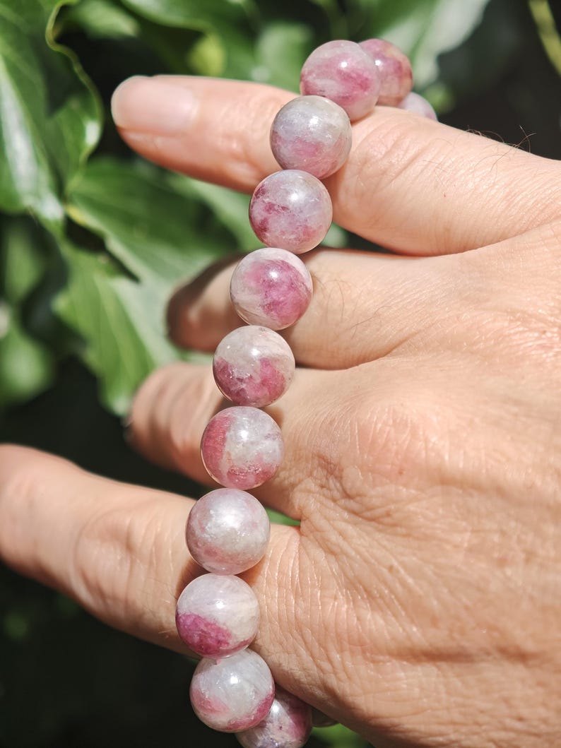 May include: A beaded bracelet featuring smooth, spherical stones with a marbled appearance in shades of deep pink, magenta, and translucent white. The beads are strung together in a single row, showcasing natural color variations and polished surfaces.
