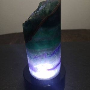 May include: A green, purple, and white fluorite crystal lamp with a black base. The crystal is illuminated from within, creating a soft glow.