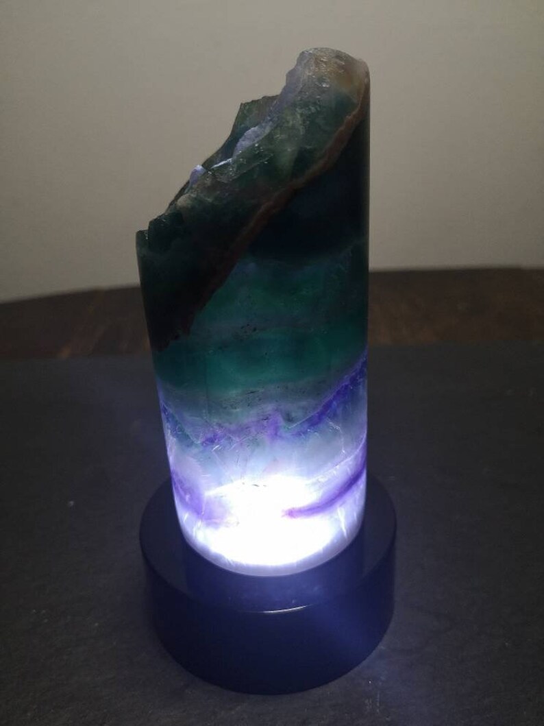 May include: A green, purple, and white fluorite crystal lamp with a black base. The crystal is illuminated from within, creating a soft glow.