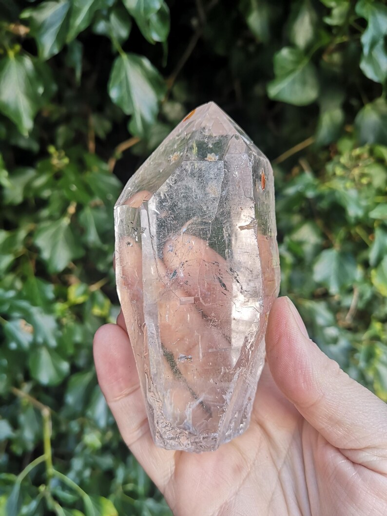 May include: A clear quartz crystal point with a natural, raw finish. The crystal is held in a hand, showcasing its size and shape.